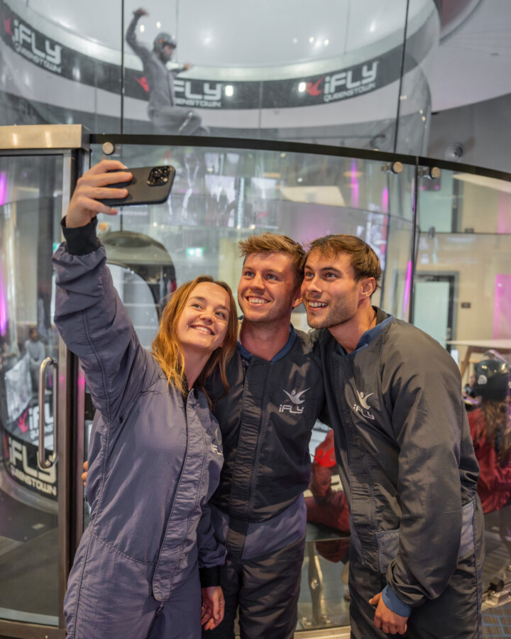 Group of friends in their mid-20's taking a selfie in front of the iFLY wind tunnel. An instructor performing tricks can be seen in the background.